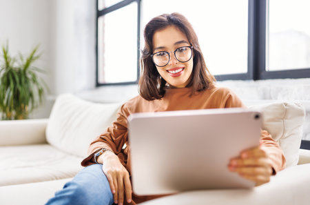 Happy young woman spending time with her pet beloved dog at home sitting on couch and browsing news in tabletの写真素材