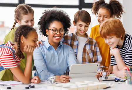 Happy female tutor and optimistic children smiling and watching tablet screen while gathering around table during lesson at schoolの写真素材