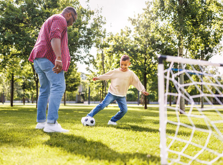 Happy african american family father and son playing football in the park in summerの写真素材