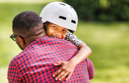 Happy african american boy in helmet hugging dad after cycling together in parkの写真素材
