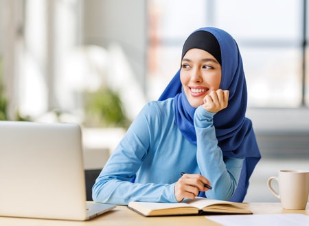 beautiful young smiling muslim woman in traditional religious hijab works remotely on laptop from homeの写真素材