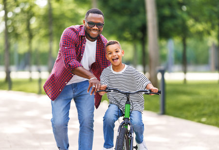 Happy african american family father teaches boy son to ride bike in park in natureの写真素材