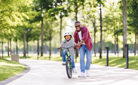 Happy african american family father teaches boy son to ride bike in park in natureの写真素材