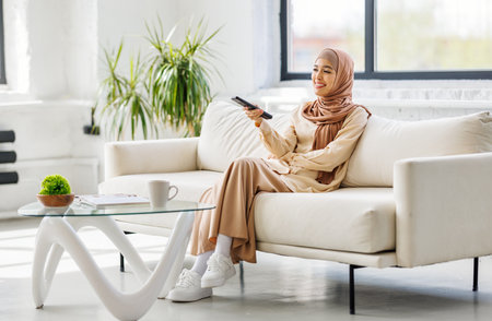 Cheerful muslim woman in hijab holding tv remote and watching series on sofa at homeの写真素材
