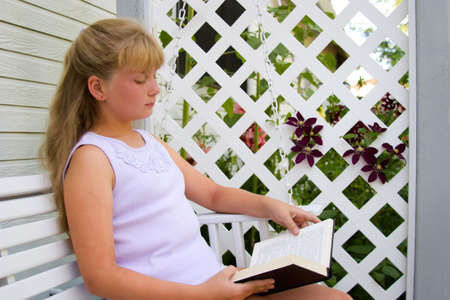 Cute blond girl reading book in shade of arbor outside her homeの写真素材
