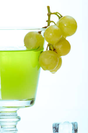 Glass of green colored liquor, bunch of grapes dipped into it with melted ice cubes around over white background with aqua colored gelの写真素材