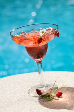 Glass of Strawberry Summer alcohol cocktail garnished with wild strawberries on swimming pool side outside of hotel in hot day, suited for vacation or travel themeの写真素材