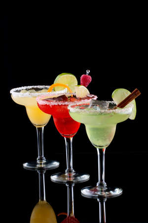 Three Margaritas - apple, orange and raspberry - in chilled glasses over black background, garnished with slice of green apple, limes, orange twist, raspberry and cinnamon stick. Most popular cocktails series.の写真素材