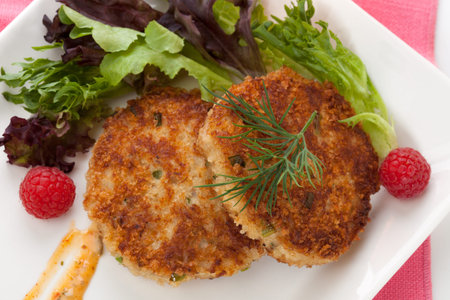 Two crab cakes appetizer garnished with spicy sauce, green salad, and raspbery 