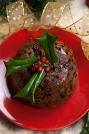 Holiday table with Christmas pudding decorated with holly twig, glass of brandy, ornaments, candles, and xmas tree の写真素材