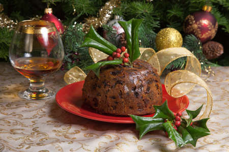 Holiday table with Christmas pudding decorated with holly twig, glass of brandy, ornaments, candles, and xmas tree の写真素材