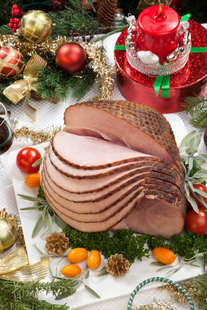 Christmas dinning table with glazed roasted ham with tomatoes, herbs, and kumquats. Surrounded by Christmas ornaments, gifts, candles, and glasses of red wine. Christmas tree in background.の写真素材