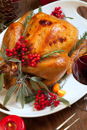 Roasted turkey garnished with sage, rosemary, and red berries in a tray prepared for Christmas dinner. Holiday table, candles and Christmas tree with ornaments.の写真素材