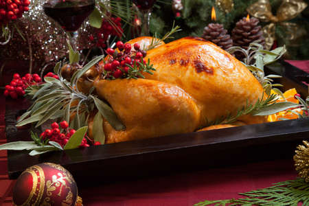 Roasted turkey garnished with sage, rosemary, and red berries in a wooden tray on Christmas decorated table. Candles and Christmas tree with ornaments.の写真素材