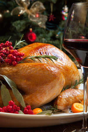 Roasted turkey garnished with sage, rosemary, and red berries in a tray prepared for Christmas dinner. Holiday table, candles and Christmas tree with ornaments.の写真素材