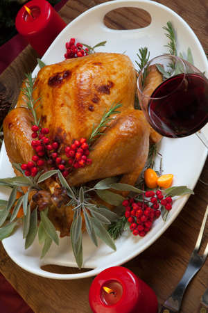 Roasted turkey garnished with sage, rosemary, and red berries in a tray prepared for Christmas dinner. Holiday table, candles and Christmas tree with ornaments.の写真素材