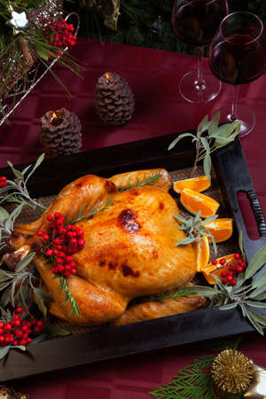 Roasted turkey garnished with sage, rosemary, and red berries in a wooden tray on Christmas decorated table. Candles and Christmas tree with ornaments.の写真素材