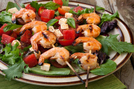 Plate of a grilled shrimp salad with feta cheese, tomatoes, and watermelon in a rustic setting.の写真素材
