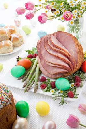 Whole baked honey sliced ham with fresh raspberry, asparagus, dyed Ester eggs, Easter cake, and cross buns. Spring flowers.の写真素材