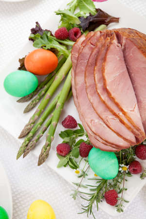 Whole baked honey sliced ham with fresh raspberry, asparagus, dyed Ester eggs, Easter cake, and cross buns. Spring flowers.の写真素材