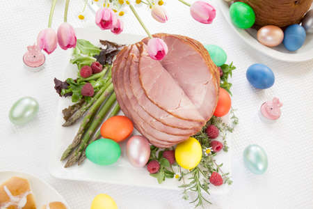 Whole baked honey sliced ham with fresh raspberry, asparagus, dyed Ester eggs, Easter cake, and cross buns. Spring flowers.の写真素材