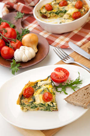 Closeup of plate with one piece of fresh made frittata, bread, and tomatoes. Pan with frittata with baby kale, sundried tomatoes, and goat cheese in background.の写真素材