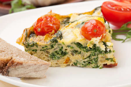 Closeup of plate with one piece of fresh made frittata, bread, and tomatoes. Pan with frittata with baby kale, sundried tomatoes, and goat cheese in background.の写真素材