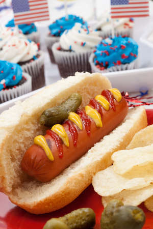Hot dog, chips, and pickled cucumbers. Angel food fruit cakes and muffins on 4th of July in patriotic theme in background.の写真素材
