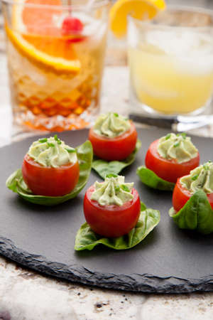 Closeup of avocado cream cheese stuffed campari tomatoes on basil leaves. Couple of tropical cocktail drinks in background.の写真素材