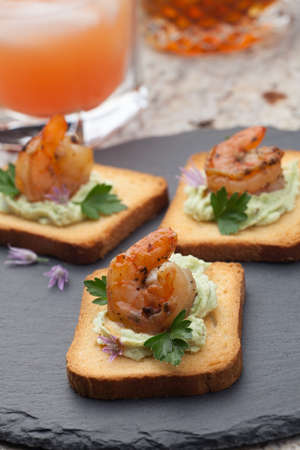 Closeup of toasts with grilled shrimps and spicy avocado spread. Couple of tropical cocktail drinks in background.の写真素材