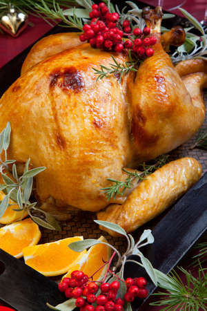 Roasted turkey garnished with sage, rosemary, and red berries in a tray prepared for Christmas dinner. Holiday table, candles and Christmas tree with ornaments.の写真素材