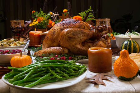 Roasted turkey garnished with cranberries on a rustic style table decoraded with pumpkins, gourds, asparagus, brussel sprouts, baked vegetables, pie, flowers, and candles.