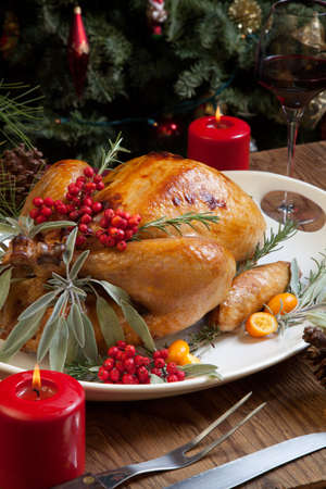 Roasted turkey garnished with sage, rosemary, and red berries in a tray prepared for Christmas dinner. Holiday table, candles and Christmas tree with ornaments.の写真素材