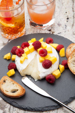 Warm baked Brie cheese with fresh mango and raspberries - great snack for cocktail party. Couple of cocktails in background.の写真素材