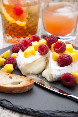 Warm baked Brie cheese with fresh mango and raspberries - great snack for cocktail party. Couple of cocktails in background.の写真素材