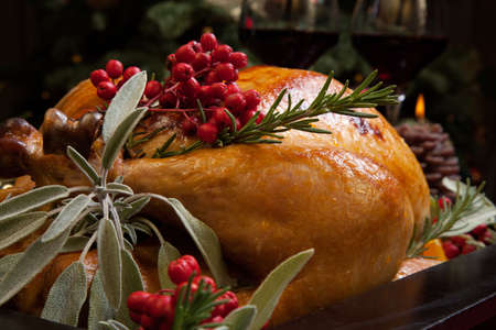 Roasted turkey garnished with sage, rosemary, and red berries in a tray prepared for Christmas dinner. Holiday table, candles and Christmas tree with ornaments.の写真素材
