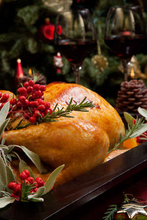 Roasted turkey garnished with sage, rosemary, and red berries in a tray prepared for Christmas dinner. Holiday table, candles and Christmas tree with ornaments.の写真素材