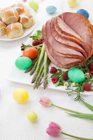 Whole baked honey sliced ham with fresh raspberry, asparagus, dyed Ester eggs, Easter cake, and cross buns. Spring flowers.の写真素材