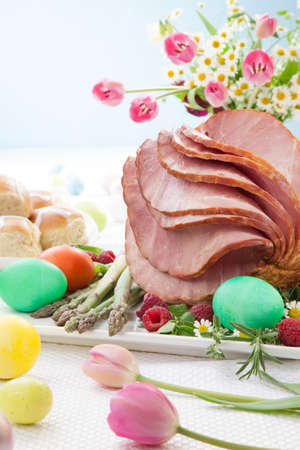Whole baked honey sliced ham with fresh raspberry, asparagus, dyed Ester eggs, Easter cake, and cross buns. Spring flowers.の写真素材