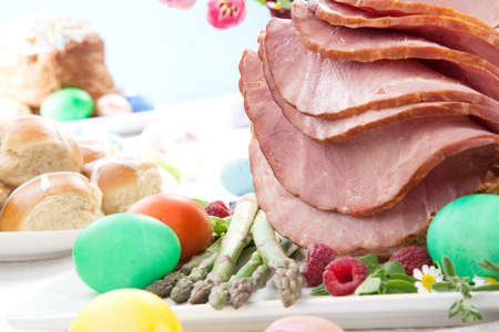 Whole baked honey sliced ham with fresh raspberry, asparagus, dyed Ester eggs, Easter cake, and cross buns. Spring flowers.の写真素材