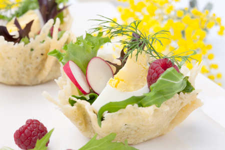 Parmesan baskets filled with fresh garden salad, hard-boiled eggs, and fresh raspberries. Easter decorated eggs, spring flowers, holiday theme. Over white.の写真素材