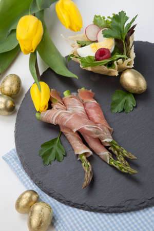 Grilled asparagus wrapped in prosciutto and parmesan baskets filled with fresh garden salad. Easter decorated eggs, tulips flowers, holiday theme.の写真素材