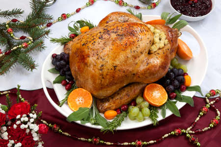 Roasted herb rubbed turkey garnished with fresh grapes, oranges, and cranberry is ready for Christmas dinner. Ornaments, Champagne, candles, and other Christmas decorations on feast table.の写真素材