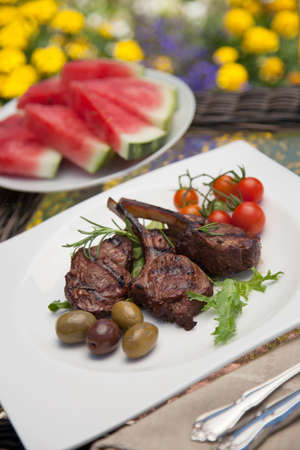 Grilled lamb chops served with cherry tomatoes and olives. Watermelon and wine. Outside dinner in a garden.の写真素材