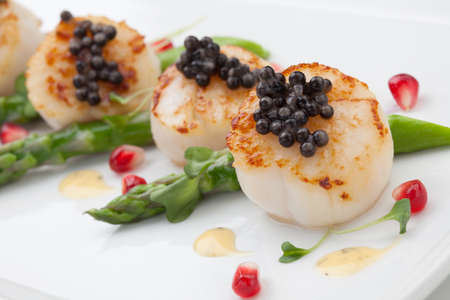 Close up of delicious appetizer scallops with black caviar. Garnished with asparagus, pomegranate seeds, microgreens, and sauce. Elite food.の写真素材
