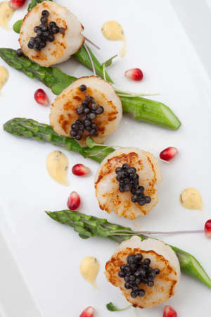 Close up of delicious appetizer scallops with black caviar. Garnished with asparagus, pomegranate seeds, microgreens, and sauce. Elite food.の写真素材