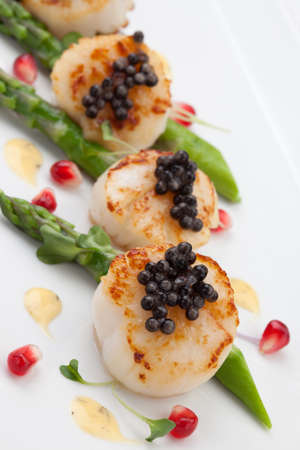 Close up of delicious appetizer scallops with black caviar. Garnished with asparagus, pomegranate seeds, microgreens, and sauce. Elite food.の写真素材