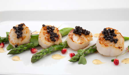 Close up of delicious appetizer scallops with black caviar. Garnished with asparagus, pomegranate seeds, microgreens, and sauce. Elite food.の写真素材