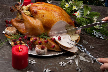 Carving rustic style roasted Christmas turkey garnished with roasted garlic, lemon, and rosehips. Surrounded with rustic Christmas ornaments, candles, wine, flowers, and Christmas tree in the background.の写真素材