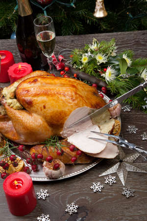 Carving rustic style roasted Christmas turkey garnished with roasted garlic, lemon, and rosehips. Surrounded with rustic Christmas ornaments, candles, wine, flowers, and Christmas tree in the background.の写真素材